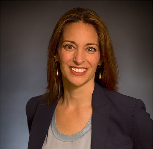 A person with long brown hair, wearing a navy blazer and light blue top, smiles against a dark background.