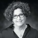 Person with curly hair and glasses wearing a dark shirt and necklace, smiling at the camera.