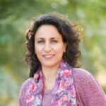 A person with curly hair and a floral scarf stands outdoors, smiling. Green foliage is blurred in the background.