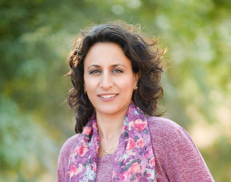 A person with curly hair and a floral scarf stands outdoors, smiling. Green foliage is blurred in the background.