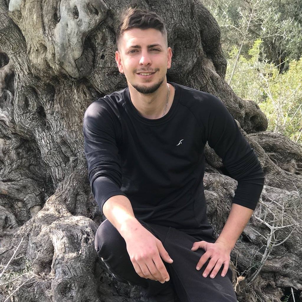 A person wearing a black long-sleeve shirt is sitting outdoors, leaning against a large, textured tree trunk.